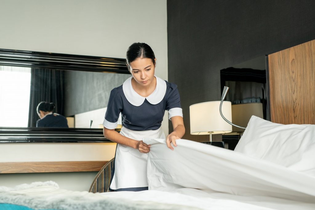 Young pretty brunette maid putting white clean blanket while changing bed in one of hotel rooms